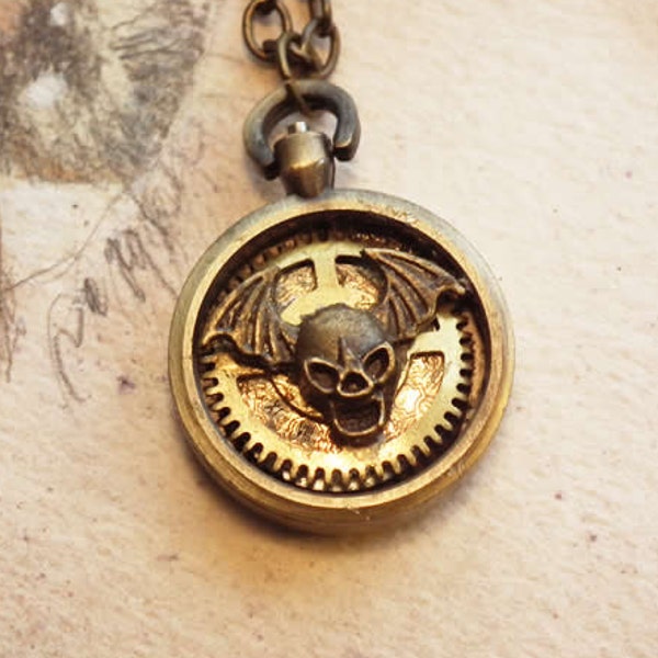 Pendentif steampunk-gothique, petit boitier de montre gousset orné d'un crane aux ailes de chauve-souris, de rouages, de strass q-09i-011