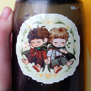 Op de afbeelding: Een sticker met twee cartoonfiguren met gesloten ogen. De figuren dragen bruine en groene kleding en zijn omringd door groene bladeren en gele harten.