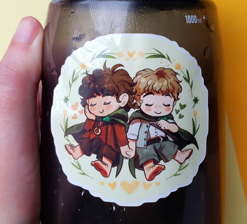 Op de afbeelding: Een sticker met twee cartoonfiguren met gesloten ogen. De figuren dragen bruine en groene kleding en zijn omringd door groene bladeren en gele harten.