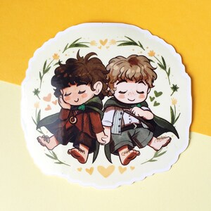 Op de afbeelding: Een sticker met twee cartoonfiguren, een met bruin haar en een groene mantel, de ander met blond haar en een groene mantel, die hand in hand staan. Ze worden omringd door gele bloemen en harten op een witte achtergrond.