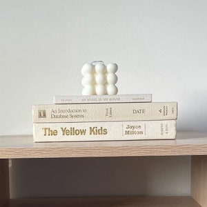 May include: A light wood bedside table with a woven rattan drawer front and a shelf. Three books are stacked on top of the bedside table, including "The Yellow Kids" and "An Introduction to Database Systems". A white candle sits on top of the books.