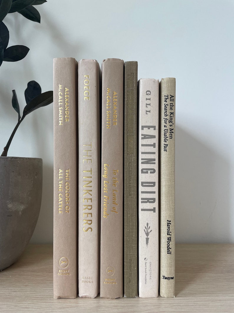 May include: A selection of hardback books with beige and olive green covers. Titles include "The Colors of All the Cattle," "The Tinkerers," and "Eating Dirt." Gold lettering and decorative elements are on the spines. A plant in a gray pot is in the background.