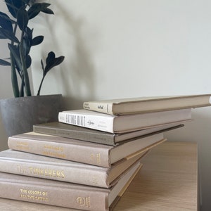 May include: A collection of hardback books in beige and grey tones, featuring gold lettering on the spines. The books are arranged on a light wood surface, with a potted plant in the background.