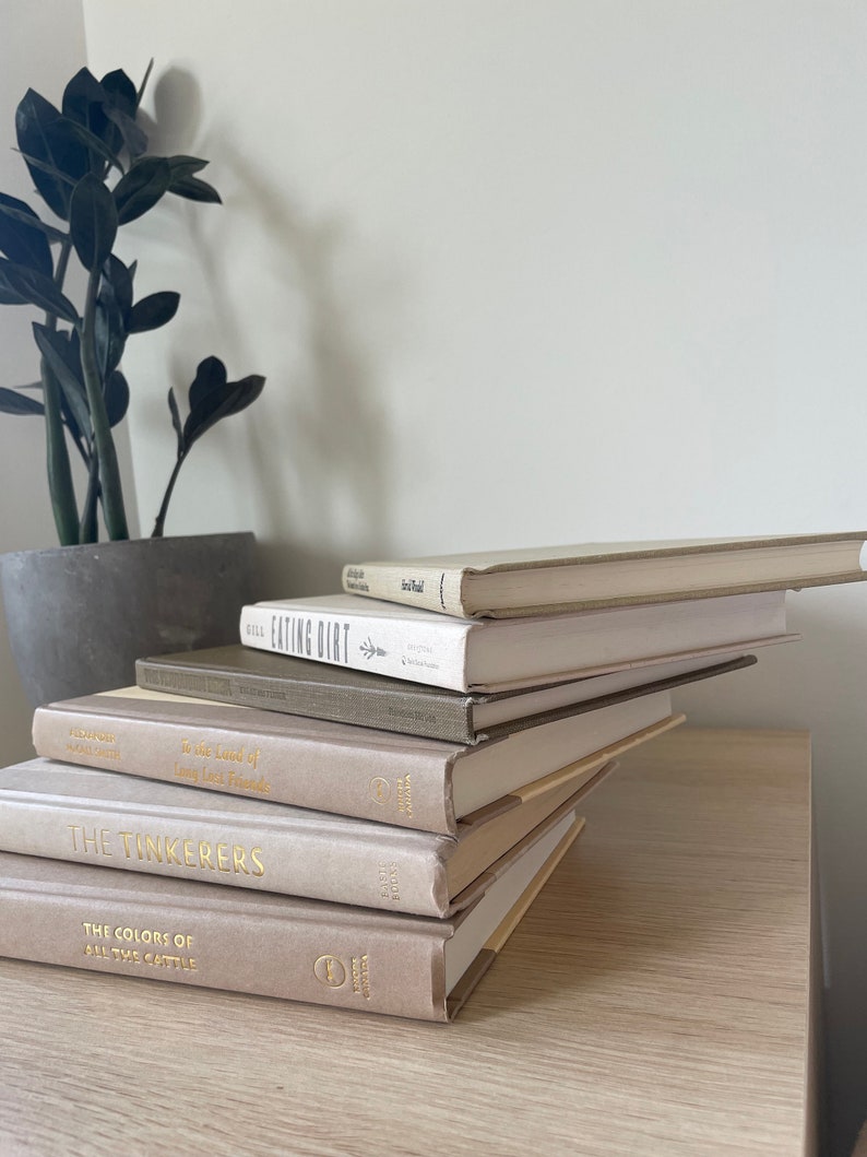 May include: A collection of hardback books in beige and grey tones, featuring gold lettering on the spines. The books are arranged on a light wood surface, with a potted plant in the background.