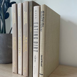 May include: A selection of hardcover books in neutral tones, including beige and grey, displayed on a light wooden shelf. The spines feature titles such as "Eating Dirt" and "All the King's Men". A potted plant is partially visible in the background.