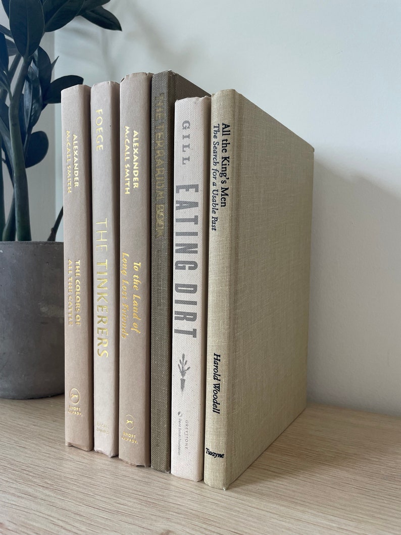 May include: A selection of hardcover books in neutral tones, including beige and grey, displayed on a light wooden shelf. The spines feature titles such as "Eating Dirt" and "All the King's Men". A potted plant is partially visible in the background.