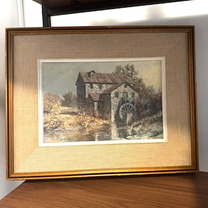 May include: A framed landscape painting of a stone mill with a water wheel, positioned beside a river. The artwork is presented in a gold-toned wooden frame with a beige linen mount. The painting shows a rural scene with trees and foliage.