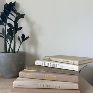 May include: A collection of books in muted tones, featuring titles such as "EATING DIRT" and "THE TINKERERS" with gold lettering. A potted plant with dark green leaves is on the left. The books are on a light-coloured wooden surface.