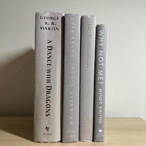 Op de afbeelding: Vier hardcover boeken met lichtgrijze covers staan op een houten oppervlak. De titels en auteurs zijn zichtbaar: "A Dance with Dragons" van George R.R. Martin, "Studio Saint-Ex" van Ania Szado, "The Postmistress" van Sarah Blake en "Why Not Me?" van Mindy Kaling.