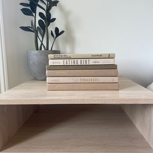 May include: A collection of books, including "Eating Dirt" and "The Tinkerers", are stacked on a light wood bedside table. A potted plant with dark green leaves is to the left. The nightstand has a lower shelf and a woven rattan door.
