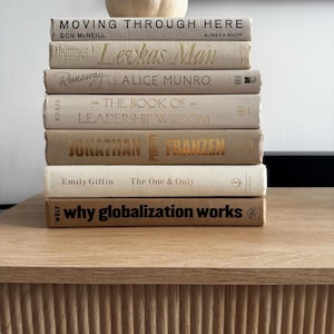 Könnte beinhalten: Ein Stapel gebundener Bücher in neutralen Farbtönen, darunter Titel wie "Moving Through Here" und "Levkas Man." Ein kleiner, cremefarbener Kürbis sitzt oben auf den Büchern. Die Bücher sind auf einer hellen Holzoberfläche angeordnet.