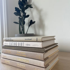 May include: A collection of hardback books in muted colours, including titles such as "Eating Dirt" and "The Tinkerers." The books are stacked on a light surface, with a potted plant in the background. The spines have gold lettering and different textures.