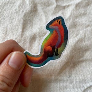 Peut inclure: Un autocollant coloré représentant un renard stylisé. Le renard est représenté dans des tons d'orange, de rouge et de violet, avec une queue arc-en-ciel. L'autocollant a une bordure blanche.