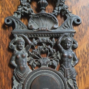 May include: Ornate bronze door hardware featuring a keyhole and two sculpted figures. The design includes floral and foliate elements, set against a wood grain background. A decorative piece for doors.