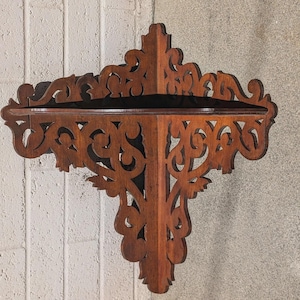 May include: A dark brown, corner wall shelf with intricate, openwork scroll designs. The shelf has two levels and is made of wood. The shelf is mounted on a light-colored, textured wall.