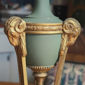 May include: A decorative, antique-style vase in muted green with gold accents. The vase features ram's head details on the sides and ornate gold trim. The base and stem are also gold, creating a classic, elegant look.