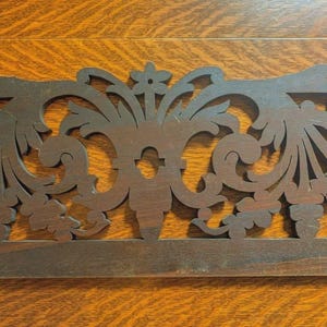 May include: A dark brown, intricately carved wooden decorative piece. The design features floral and scrollwork patterns, with a scalloped top edge and a flat base. The wood has a natural grain and a matte finish, suggesting an antique or vintage aesthetic.
