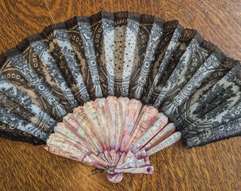 May include: An open antique fan with a mother-of-pearl base and patterned fabric panels. The fabric features a black and white floral design with a lace trim. The fan is resting on a wooden surface.