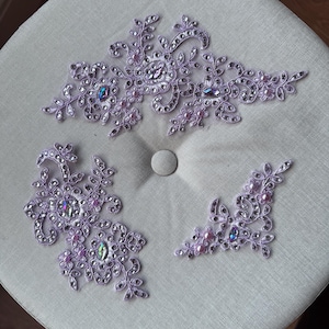 May include: Three lavender lace appliques with floral and leaf designs. Each applique is embellished with clear rhinestones and iridescent gems. Small pink pearls are also incorporated into the design. The appliques are arranged on a light grey surface.
