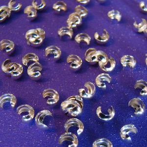 May include: A close-up of a group of silver-toned metal crimp beads. The beads are scattered on a purple surface.