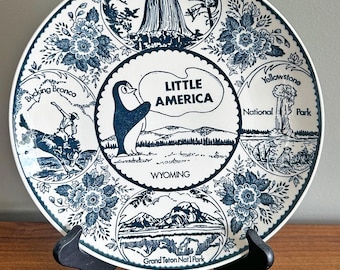 May include: A round white ceramic plate with dark blue illustrations featuring a central penguin and the text Little America Wyoming. Surrounding scenes depict Devils Tower, a bucking bronco, Yellowstone National Park, and Grand Teton Nat'l Park.