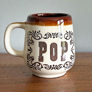 May include: Cream-colored ceramic mug with a brown rim and handle. The word "POP" is printed in large, brown letters on the front, surrounded by decorative scrollwork. The mug is sitting on a wooden surface.