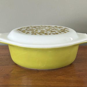 May include: A vintage, lime green Pyrex casserole dish with a white lid. The lid features a pattern of gold-colored floral designs. The dish has a rectangular shape with rounded corners and handles. A classic piece of kitchenware.
