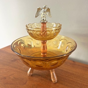 May include: A two-tiered amber glass serving dish with a wooden center post and three wooden legs. The top tier is smaller than the bottom tier. A gold-colored eagle tops the center post. The dish has a decorative pattern.