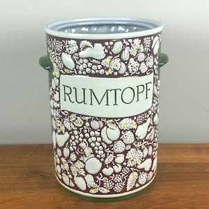 May include: A cylindrical ceramic container with the word "RUMTOPF" in green lettering. The container is decorated with a pattern of white and yellow fruits and leaves on a dark maroon background. It has two green handles.