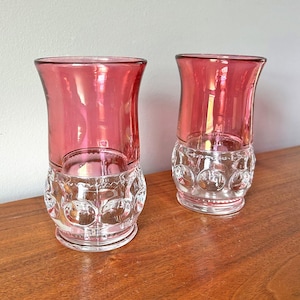 May include: Two vintage pink glass tumblers. The upper portion of each glass is a vibrant pink, while the lower portion features a clear, patterned design. The glasses are displayed on a wooden surface, showcasing their elegant design.