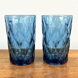 May include: Two blue drinking glasses with a diamond pattern. The glasses are made of a transparent material and are sitting on a wooden surface. The glasses are empty and have a simple, elegant design.