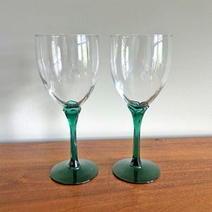 May include: Two clear wine glasses with green stems and bases. The stems have a decorative, twisted design. The glasses are empty and sit on a wooden surface.