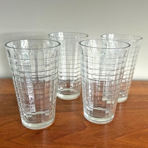 May include: Four clear, tall drinking glasses with a grid pattern. The glasses are cylindrical with a slightly flared rim and sit on a wooden surface. The glasses are empty and appear to be made of glass.