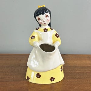 May include: A vintage ceramic figurine of a girl in a yellow dress with brown flower accents and a white apron. The figurine has black hair, a yellow bow, and is holding a container. The piece is likely a decorative vase or planter.