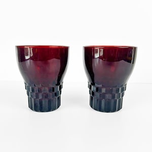 Peut inclure: Deux verres à boire de couleur bordeaux. Les verres ont un dessus arrondi et un design géométrique à gradins à la base. Les verres sont en matériau brillant et sont posés sur un fond blanc.