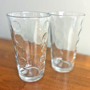May include: Two clear drinking glasses with a textured, bubble-like pattern. The glasses are empty and sit on a wooden surface. The glasses have a cylindrical shape with a slightly flared top.