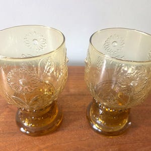 May include: Two amber-colored, stemmed glasses. The glasses have a rounded bowl shape with a floral pattern and a decorative base. The glasses are made of glass and are sitting on a wooden surface.