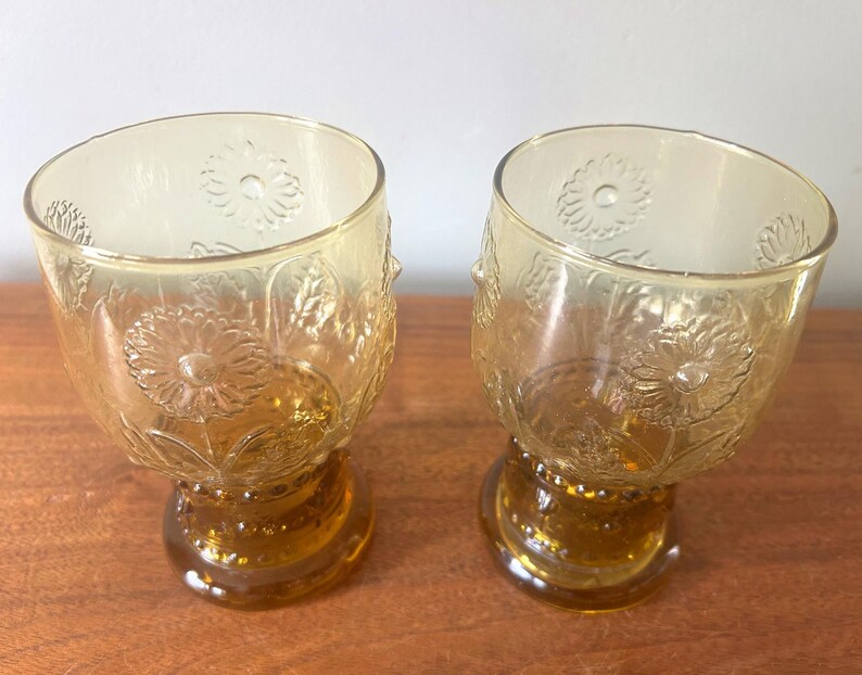 May include: Two amber-colored, stemmed glasses. The glasses have a rounded bowl shape with a floral pattern and a decorative base. The glasses are made of glass and are sitting on a wooden surface.