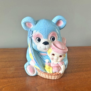 May include: A blue ceramic piggy bank shaped like a bear holding a doll with a pink hat and dress. The bear has a yellow and blue striped scarf and the doll is sitting in a wicker basket.