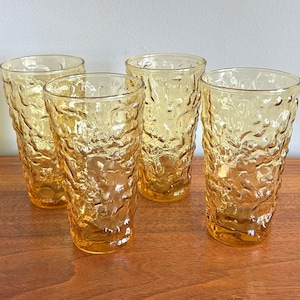 May include: A set of four amber-colored glass tumblers featuring a textured, bumpy surface pattern. Each cylindrical drinking glass has a thick base and a translucent, golden-yellow hue that catches the light, creating a distinct visual depth.