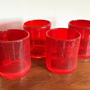 May include: A set of four cylindrical drinking glasses made of vibrant red glass. Each glass features a distinct bubbled texture throughout the material, with a smooth rim and a flat base, arranged in a group of four on a flat surface.