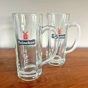 May include: Two clear glass beer mugs with handles. Each mug has the Heineken logo in green, red, and white. The mugs are tall and rectangular, perfect for enjoying a cold beverage.