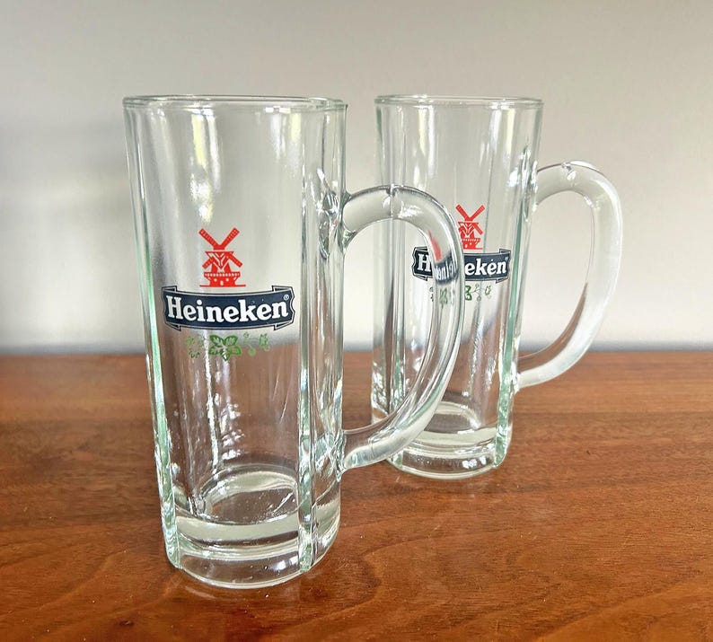 May include: Two clear glass beer mugs with handles. Each mug has the Heineken logo in green, red, and white. The mugs are tall and rectangular, perfect for enjoying a cold beverage.