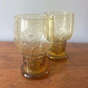 May include: Two amber-colored vintage drinking glasses. The glasses have a floral design etched into the glass. The base of the glasses are a darker amber color and have a decorative beaded design. The glasses are likely made of glass.