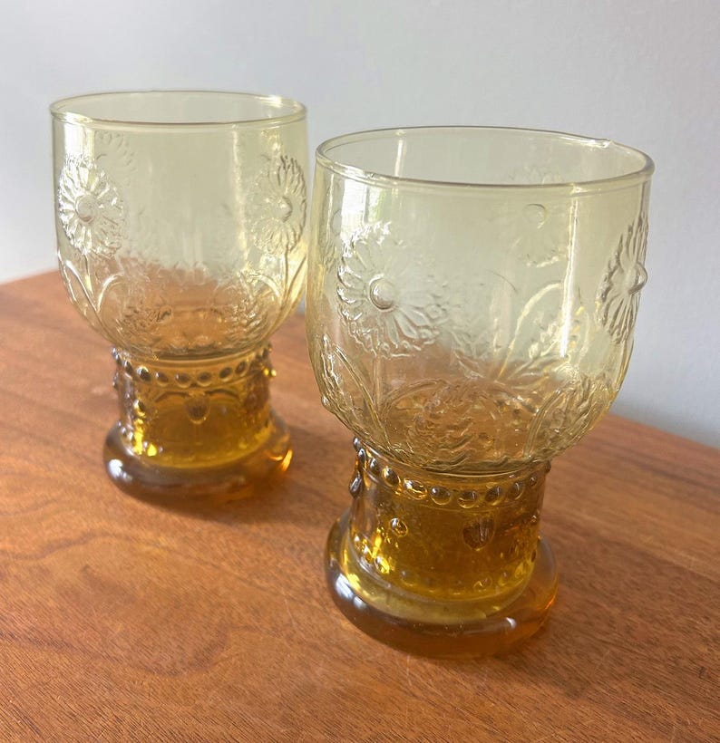 May include: Two amber-colored vintage drinking glasses. Each glass features a floral design etched into the glass. The base of each glass is a darker amber color, with a decorative pattern. The glasses are on a wooden surface.