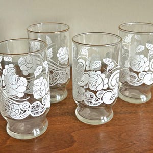 May include: Four clear drinking glasses with a white floral design. The glasses have a gold rim and a decorative white floral pattern. The glasses are on a wooden surface.