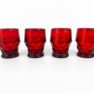 May include: Four ruby red drinking glasses. Each glass has a faceted design on the lower half and a smooth rim. The glasses are evenly spaced and sit on a white surface. The glasses are a vintage style.