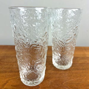 May include: Two clear, textured drinking glasses. The glasses have a tall, cylindrical shape with a textured surface. The texture gives the glasses a unique, organic appearance. They are set on a wooden surface.
