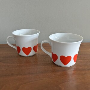 May include: Two white ceramic coffee mugs with red heart designs. The mugs have a simple, rounded shape and a handle on the side.