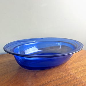 May include: A vibrant blue glass bowl with a wide, shallow design. The bowl has a smooth, glossy finish and sits on a wooden surface. The bowl's shape is oval, with a slightly raised rim.